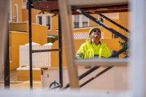 Positiv gelernter grauhaariger Arbeiter in Uniform und Schutzhelm, der bei sonnigem Wetter hinter einer Metallkonstruktion an einer Hauswand steht und wegschaut. — Stockfoto