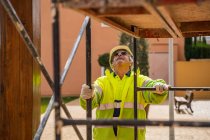 Positive skilled gray haired workman in uniform and protective helmet looking away while standing behind metal construction against building wall in sunny day — Stock Photo