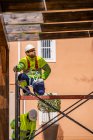 Professional engineer in uniform and helmet sitting on scaffolding and looking away while working with colleague on maintenance of electric equipment near residential building — Stock Photo