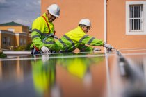 Group of workers in uniform and helmets installing photovoltaic panels on roof of wooden construction near house — Stock Photo