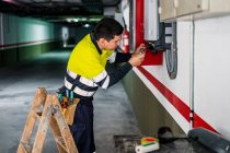 Seitenansicht eines jungen professionellen männlichen Technikers mit elektrischen Werkzeugen, der Ausrüstung repariert und überprüft, während er im Gebäude arbeitet — Stockfoto
