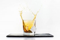 Glass of yellow cold drink with falling ice cube and splashes placed on black slate board on white background — Stock Photo