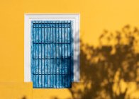 Fenster mit schäbigen blauen Holzläden an leuchtend gelber Hauswand im Sonnenschein schließen — Stockfoto