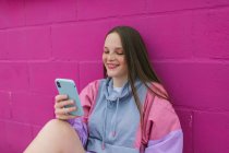 Adolescente na moda sentado perto da parede rosa com smartphone — Fotografia de Stock