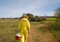 Обратный вид на неузнаваемого пчеловода в профессиональном желтом костюме, несущего пластиковый контейнер во время прогулки по зеленому полю в солнечный летний день — стоковое фото