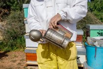 Primer plano de la cosecha apicultor masculino anónimo haciendo fuego en el fumador de abejas mientras trabaja en el colmenar - foto de stock