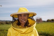Положительная женщина-пчеловод в желтом защитном костюме и маске улыбается и смотрит в камеру, стоя на зеленом поле на пасеке в солнечный летний день — стоковое фото