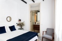 Удобная кровать с бело-синим пододеяльником и мягким креслом, расположенная у дверей ванной комнаты в уютной спальне современной квартиры — стоковое фото