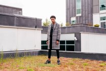 Serious jovem afro-americano elegante cara na moda casaco xadrez e calças apertadas olhando para a câmera, enquanto de pé contra edifícios modernos na cidade — Fotografia de Stock