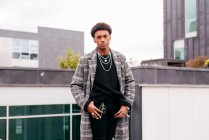 Seriöser junger afroamerikanischer Modemacher im trendigen karierten Mantel und engen Hosen blickt in die Kamera, während er vor modernen Gebäuden in der Stadt steht — Stockfoto