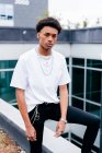 Молодий афроамериканець у модний одяг, сталеві ланцюжки та браслет, який дивиться на камеру, стоячи біля сучасного будинку в місті. — стокове фото