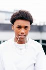Junger afroamerikanischer Typ mit verschiedenen Ketten und Ringen, Kette im Mund und Blick in die Kamera, während er vor verschwommenem modernem Gebäude steht — Stockfoto