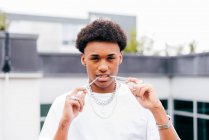Young African American guy with various chain necklaces and rings holding chain in mouth and looking at camera while standing against blurred modern building — Stock Photo