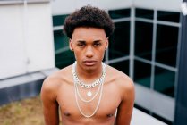 From above serious young African American male with naked torso wearing various trendy steel neck chains and pendant looking at camera while standing against modern building — Stock Photo