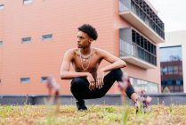 Confident young African American male with chain necklaces on naked torso dressed in tight pants and trendy boots squatting on street with modern building in background — Stock Photo