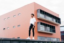 Знизу - молодий стильний афроамериканець, який дивиться у білу сорочку і щільні штани з білими кросівками, що стоять навпроти рожевого міського будинку. — стокове фото
