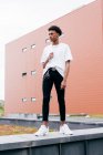 Low angle of stylish independent young African American man in trendy outfit with steel accessories looking at camera while standing against pink building on city street — Stock Photo