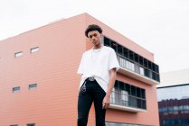 Низкий угол стильного независимого молодого афроамериканца в модном наряде со стальными аксессуарами, смотрящего в камеру, стоя напротив розового здания на городской улице — стоковое фото