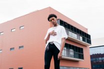 Baixo ângulo de elegante jovem afro-americano independente em roupa da moda com acessórios de aço olhando para a câmera, enquanto em pé contra edifício rosa na rua da cidade — Fotografia de Stock