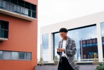 Снизу счастливый молодой афроамериканец в стильном наряде просматривает смартфон, стоя на городской улице рядом с современным зданием — стоковое фото