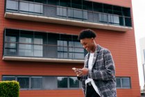 From below side view of happy young African American male student in stylish outfit browsing smartphone while standing on city street near modern building — Stock Photo