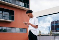 De dessous vue latérale de heureux jeune étudiant afro-américain mâle naviguant smartphone tout en marchant sur la rue de la ville près du bâtiment moderne — Photo de stock