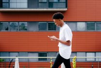 From below side view of happy young African American male student browsing smartphone while walking on city street near modern building — Stock Photo