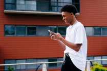 Desde abajo vista lateral de la joven estudiante afroamericana feliz navegando por el teléfono inteligente mientras camina por la calle de la ciudad cerca del edificio moderno - foto de stock