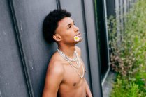 Side view alluring young black teenage man with naked torso and neck chains holding flower in mouth and looking away while standing against gray wall — Stock Photo