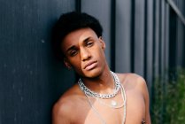 Alluring young black teenage man with naked torso and neck chains holding flower in mouth and looking at camera while standing against gray wall — Stock Photo