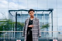 Selbstbewusstes junges afroamerikanisches Teenie-Model im trendigen Schottenmantel und stilvollen Accessoires, das vor einem modernen Gebäude mit Glaswand in die Kamera blickt — Stockfoto