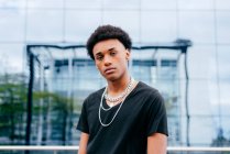 From below of young curly haired African American teenage guy in black t shirt and trendy neck chains looking at camera while standing against blurred modern building with glass wall — Stock Photo