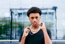 From below of young curly haired African American teenage guy in black t shirt and trendy neck chains looking at camera while standing against blurred modern building with glass wall — Stock Photo