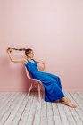 Side view of young woman in blue dress barefoot looking at camera holding ponytail while sitting in chair and looking away on minimalist pink background — Stock Photo