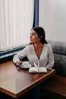 Brünette Frau mit langen Haaren trinkt Kaffee in einer Bar mit einem Buch daneben — Stockfoto