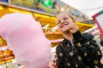 Von unten entzückte Mädchen lächeln und essen süße Zuckerwatte, während sie auf Jahrmarkt stehen — Stockfoto