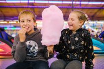 Entzückte Geschwister lächeln und essen süße Zuckerwatte, während sie auf dem Jahrmarkt sitzen — Stockfoto