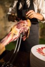 Crop unrecognizable person hand on apron cutting a whole dry-cured ham leg on a black background — Stock Photo