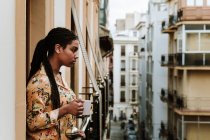 Вид сбоку спокойной молодой афроамериканки в повседневной одежде с чашкой горячего напитка, стоящей на балконе старого жилого дома в городе — стоковое фото