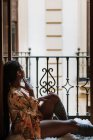 Vista laterale della silenziosa donna etnica sognante in abito casual seduta sul balcone e distogliendo lo sguardo mentre riposa a casa durante il fine settimana — Foto stock