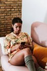 Moderna donna afroamericana in abbigliamento casual seduta su comodo divano e smartphone navigante mentre riposa in un accogliente soggiorno con interni soppalcati — Foto stock