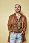 Fashionable male model with tattoos wearing trendy coat over leopard shirt and jeans standing against beige background and looking at camera with hands in pocket — Stock Photo