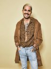 Alegre modelo masculino de moda con tatuajes con abrigo de moda sobre camisa de leopardo y jeans de pie sobre fondo beige y mirando a la cámara - foto de stock