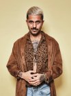 Modisches männliches Model mit Tätowierungen trägt trendigen Mantel über Leopardenhemd und Jeans vor beigem Hintergrund und blickt in die Kamera — Stockfoto
