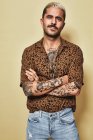 Modelo masculino elegante com tatuagens vestindo camisa de leopardo na moda e jeans em pé contra fundo bege e olhando para a câmera com os braços cruzados — Fotografia de Stock
