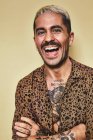 Ritratto di allegro modello maschile alla moda con tatuaggi che indossano camicia leopardata alla moda in piedi su sfondo beige e guardando la fotocamera — Foto stock