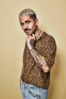 Модна чоловіча модель з татуюваннями у модній сорочці з леопардом та джинсах, що стоять на бежевому фоні та дивляться на камеру — стокове фото