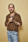 Modelo masculino elegante com tatuagens vestindo camisa de leopardo na moda e jeans em pé contra fundo bege e olhando para a câmera — Fotografia de Stock