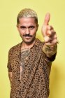 Adult bearded guy with stylish haircut and tattoo dressed in leopard shirt making finger gun gesture and looking at camera against yellow background — Stock Photo