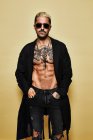 Brutal muscular sexy fit male with tattooed torso wearing black coat and trendy ripped jeans with stylish sunglasses and accessories standing against beige background — Stock Photo